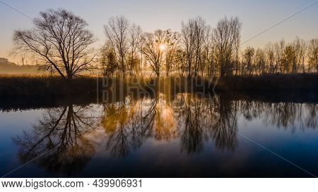 The Rays Of The Morning Sun Shining Through The Autumn Trees And Reflected In The Water Of The Misty