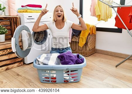 Young caucasian woman doing laundry with clothes in the basket smiling amazed and surprised and pointing up with fingers and raised arms. 