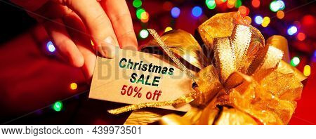 A Woman's Hand Holds A Label With Text - Christmas Sale 50 Off, On A Gift Box With A Gold Bow, Close