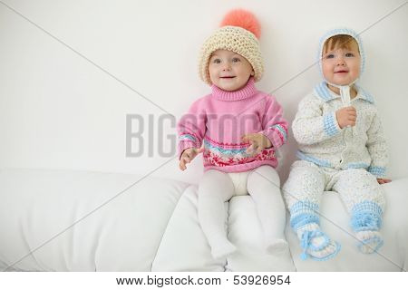 Two little kids in hats and knitted jackets sit on back of white sofa at home. Shallow depth of field.