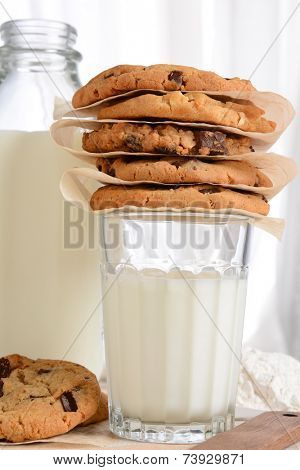 Closeup of a stack of fresh baked cookies on top of a glass of milk. Vertical format with a bottle of milk and flour in the background. Chocolate Chip, Oatmeal Raisin, White Chocolate cookies.