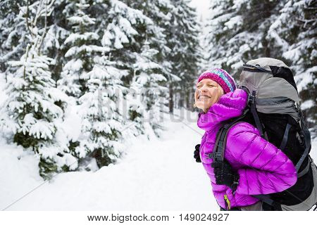 Happy young woman hiking in white winter forest woods with backpack. Girl walking on snowy trail. Inspirational travel and healthy lifestyle. Camping in snow winter landscape.