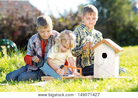 Three boys - two teenagers and one preschooler making together nesting box sitting on lawn in summerday
