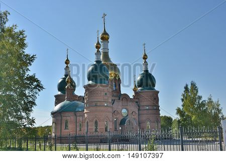Christian church with green and gold domes and crosses on them, from a red brick in the open air, behind a fence, with the bottom of the frame can be seen green grass lawn