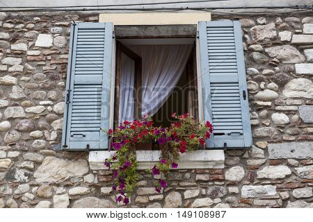 Old window with open shutters with flowers on the window sill on the stone wall. Italian Village.