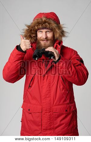 Portrait of a man wearing red winterjacket with hood on, holding binoculars and gesturing thumb up, studio shot
