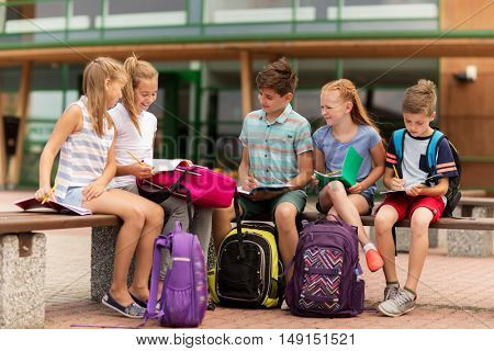 primary education, friendship, childhood, communication and people concept - group of happy elementary school students with backpacks and notebooks sitting on bench outdoors