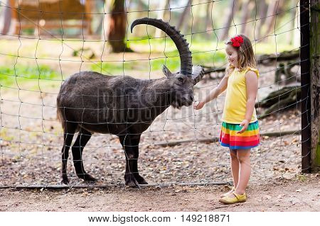 Cute little girl in colorful dress watching and feeding wild alpine goat with large horns at the zoo on sunny summer day. Wildlife and Alps mountains nature experience for kids at animal safari park.
