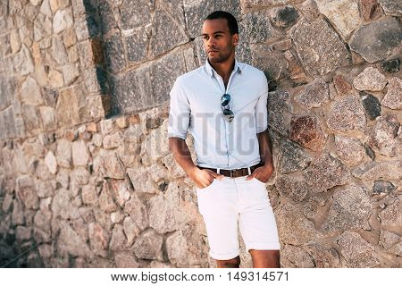 Casually handsome. Handsome young African man in smart casual clothes holding hands in pockets and looking away while standing against the stoned wall outdoors