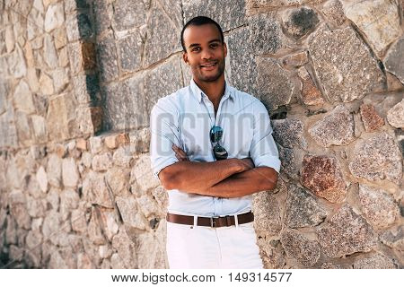 Cheerful and handsome. Handsome young African man in smart casual clothes keeping arms crossed and looking at camera with smile while standing against the stoned wall outdoors