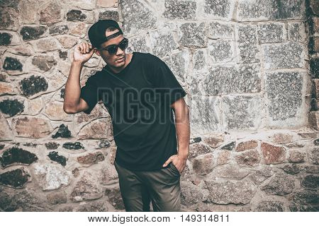 Feeling comfortable in his style. Handsome young African man in casual clothes adjusting his cap and keeping one hand in pocket while standing against the stoned wall outdoors