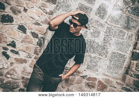 Cool and stylish. Handsome young African man in casual clothes adjusting his cap and keeping one hand in pocket while standing against the stoned wall outdoors