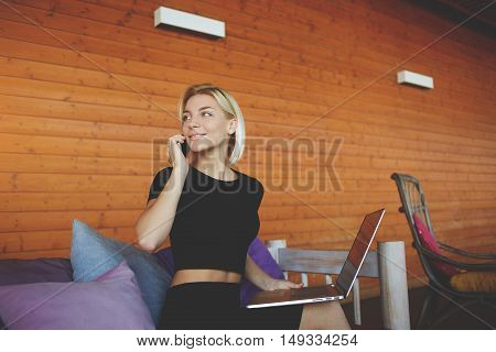 Young woman is smiling for friends during talk via smart phone while is sitting in modern co-working space.Beautiful hipster girl is calling via cell telephone during work on portable laptop computer