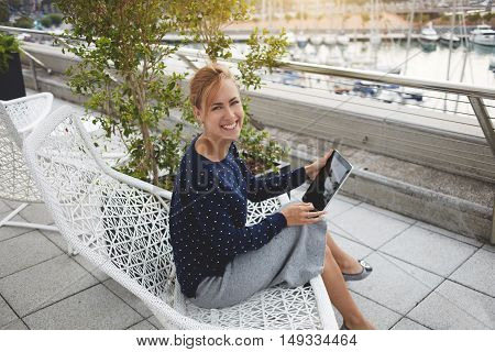 Smiling female with touch pad in hands is looking at camera while is sitting outdoors near marina port. Cheerful businesswoman is using digital tablet for reading electronic book during leisure time