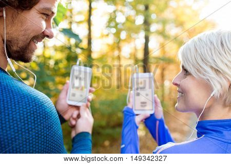 Beautiful couple running together outside in colorful sunny autumn forest using a fitness app on their smartphones. Using phone app for tracking weight loss progress, running goal or summary of their run. Rear view.