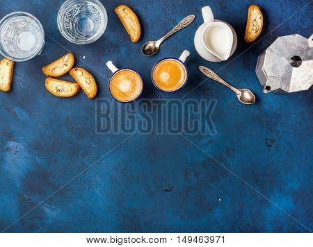 Coffee espresso in cups with italian cantucci, cookies and milk in jug over dark blue painted plywood background, top view, copy space, . Food frame concept