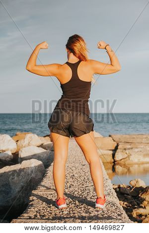 Young girl showing strong biceps after outdoors fitness workout. Fitness woman. Whole body back view.