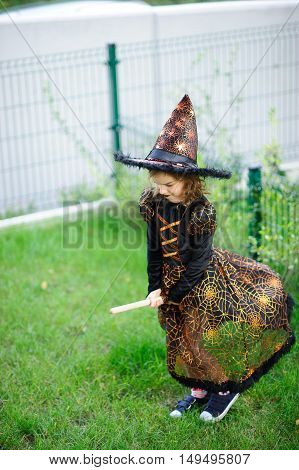 Preparation for Halloween. The girl in a suit of the evil witch pretends that she flies on the broom. The girl is dressed in a black-orange dress and a big hat. She has a good mood. Children adore Halloween.