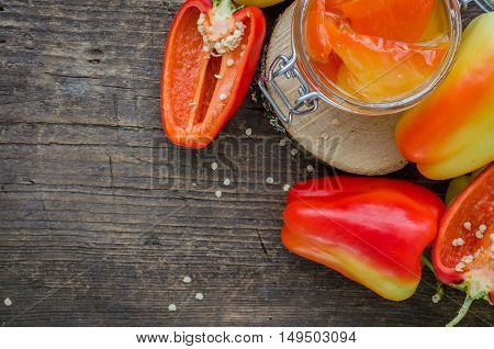 Bell pepper preserved in a glass jar with fresh peppers on old wooden background. Homemade marinated in oil red pepper. Glass jar with conserved roasted yellow and red paprika. Top view. Copy space.