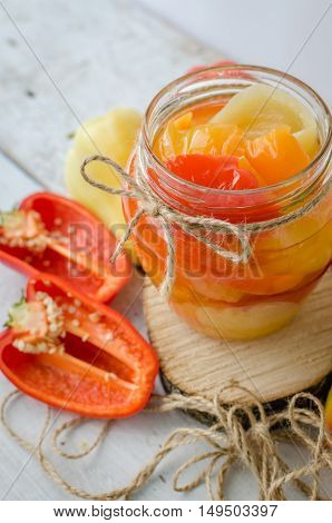 Bell pepper preserved in a glass jar with fresh peppers on white wooden background. Homemade marinated in oil red pepper. Glass jar with conserved roasted sweet paprika. Selective focus.