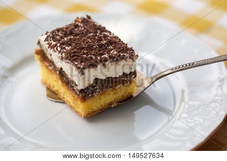 Sponge cake with cream and chocolate on slice on white plate on yellow checkered tablecloth