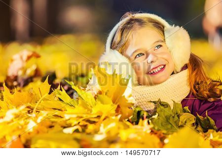 Little girl in earflaps playing with autumn leaves in the park