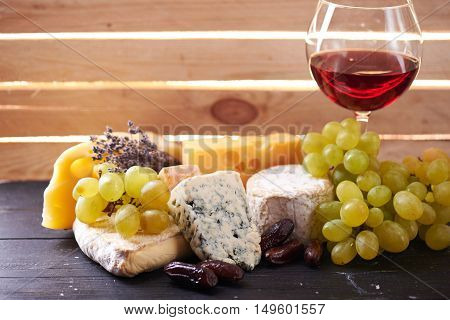 The Glass of red wine served with grapes, date fruits and the variety of cheese on a black wooden table with the wooden board on a background with a ray of light shining through it. Close view