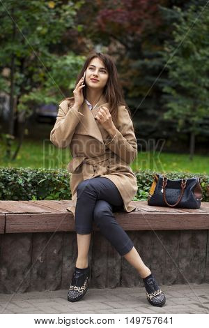 Young beautiful brunette woman in beige coat sitting on a bench in the autumn park