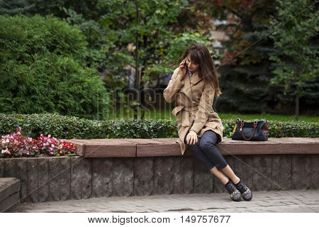 Young beautiful brunette woman in beige coat sitting on a bench in the autumn park