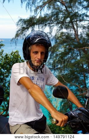 a confident young man portrait with T-shirt and helmet sitting on motorbike against sea background.