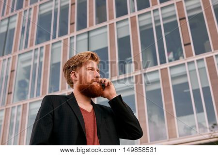 Serious and glad man talk by phone outdoor. Big glass business building background. Deal conversation on the fresh air. Proud gay in suit and red t-shirl look like hipster.