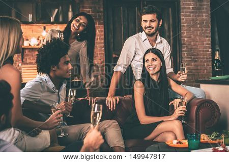 Friends gathering. Group of cheerful young people enjoying food and drinks while spending nice time in cofortable chairs at home together