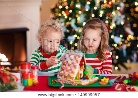 Little boy and girl making Christmas gingerbread house at fireplace in decorated living room. Kids playing with ginger bread under Christmas tree. Baking and cooking with children for Xmas at home.