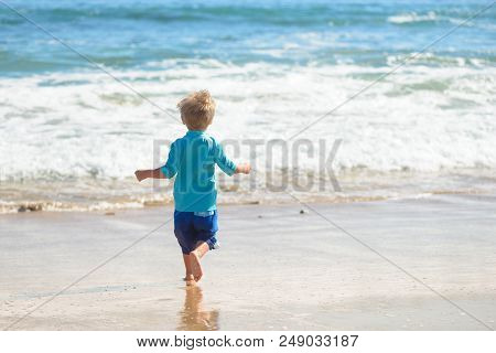 Happy Boy In Blue Swimsuit Running On The Beach In The Waves, Family On Vacation, View From Behind