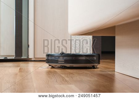 Black Robot Vacuum Cleaner Runs Under Bed On Laminate Floor In Bedroom. Modern Smart Cleaning Techno