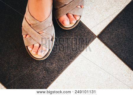 Female Feet With A Nude Pedicure In Beige Suede Sandals On The Background Of A Chessboard Floor.
