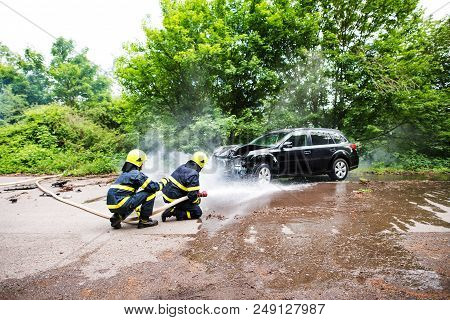 Two Firefighters Extinguishing A Burning Car After An Accident On The Road In The Countryside.