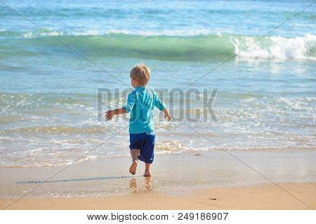 Happy Boy In Blue Swimsuit Running On The Beach In The Waves, Family On Vacation, View From Behind