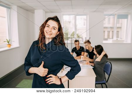 Cheerful Confident Woman Gesturing Thumb Up With Her Colleagues Working Behind Her. Friendly Atmosph