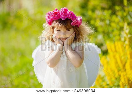 Beautiful Little Curly Blonde Angel Girl With Wreath Made Of Red Roses Smiles And Teases. Summer Day