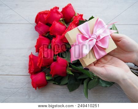 Preparing A Gift, On A Wooden Table, With Roses. Female Hand Holding A Gift, Wrapped With Ribbon, On