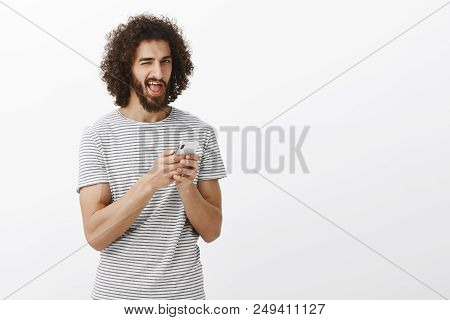 Indor Shot Of Carefree Happy Young Male Student With Beard And Afro Hairstyle, Holding Smartphone An