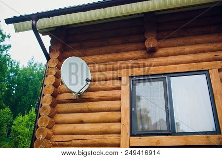 Satellite Dish On The Wall Of The House. Antenna Is White. The House Is Made Of A Wooden Beam Of Bro