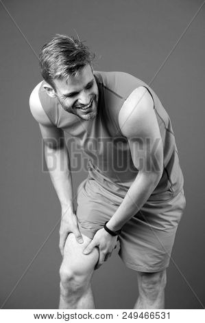 Sportsman With Knee Pain On Violet Background. Man In Orange Vest And Blue Shorts. Health Problem An