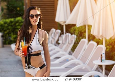 Pretty Girl With Long Dark Hair Standing By The Dack Chairs And Umrellas Near A Swimming Pool Holdin