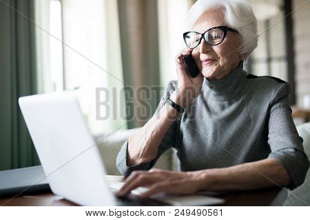 Elegant Senior Woman With White Hair Typing On Laptop During Conversation On Telephone Working Dista