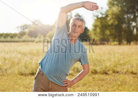Athletic Mature Male With Fit Body, Bends Aside, Does Physical Exercise Outdoor, Poses Against Green