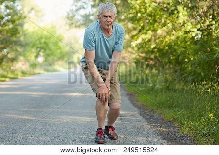 Full Length Portrait Of Displeased Male Pensioner, Leans On Knees, Has Pain After Jogging, Banged Hi