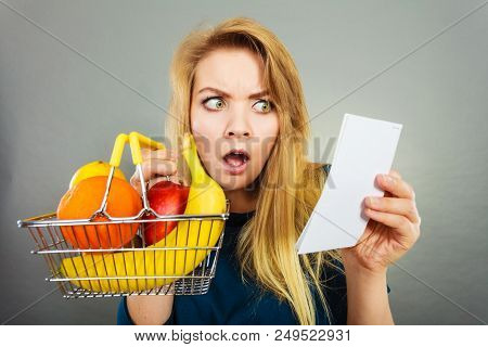 Shocked Woman Holding Shopping Basket With Fruits Looking At Bill Receipt Being Scared Of Huge Price