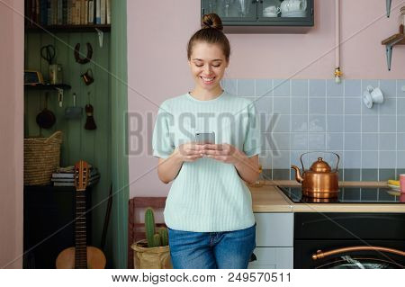 Horizontal Portrait Of Young European Woman Standing Alone On Kitchen With Smartphone In Hands Smili